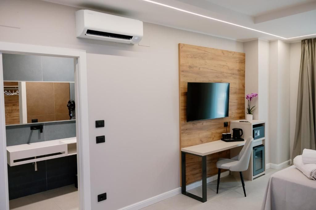 Habitación de hotel moderna con televisor de pantalla plana montado en un panel de madera, un escritorio con una silla, un mueble pequeño y una cama con cubrecama blanco.