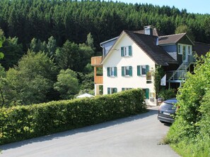 A two-story house with a balcony, a car parked in front, and a forested hill in the background. - Holiday Apartment With 1 Bedroom (Bad Berleburg)