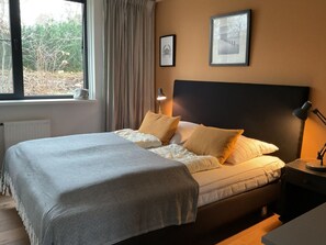 Ein Schlafzimmer mit einem Bett, einem Nachttisch, einer Lampe, einem Fenster mit Vorhängen und zwei gerahmten Bildern an der Wand.