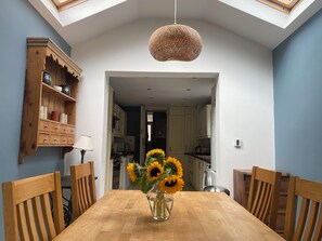 Un coin repas avec une table en bois, des chaises, un vase rempli de tournesols, un luminaire suspendu et une cuisine visible à travers une porte ouverte.