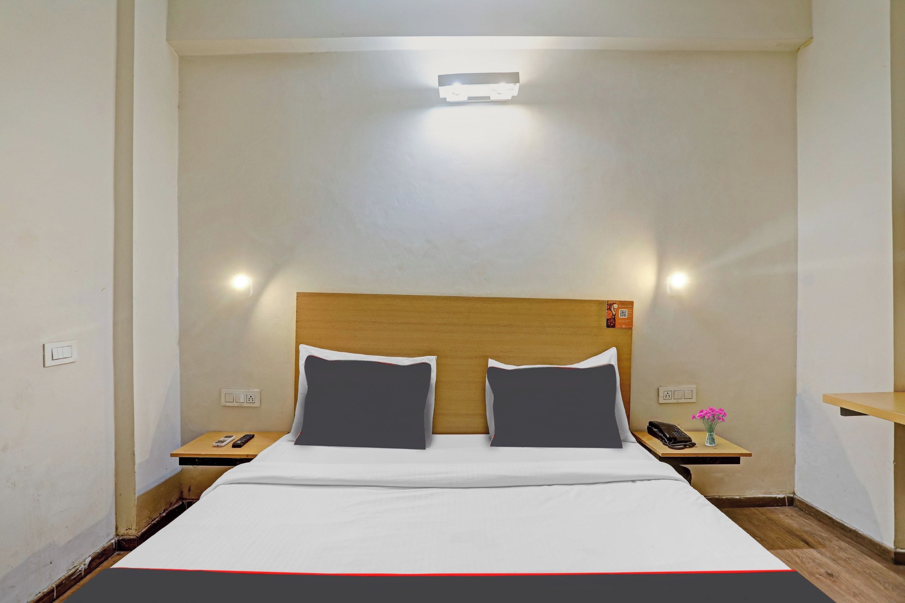A hotel room with a bed, two bedside tables, a nightstand, a wall-mounted light, and a small shelf with a vase of flowers.