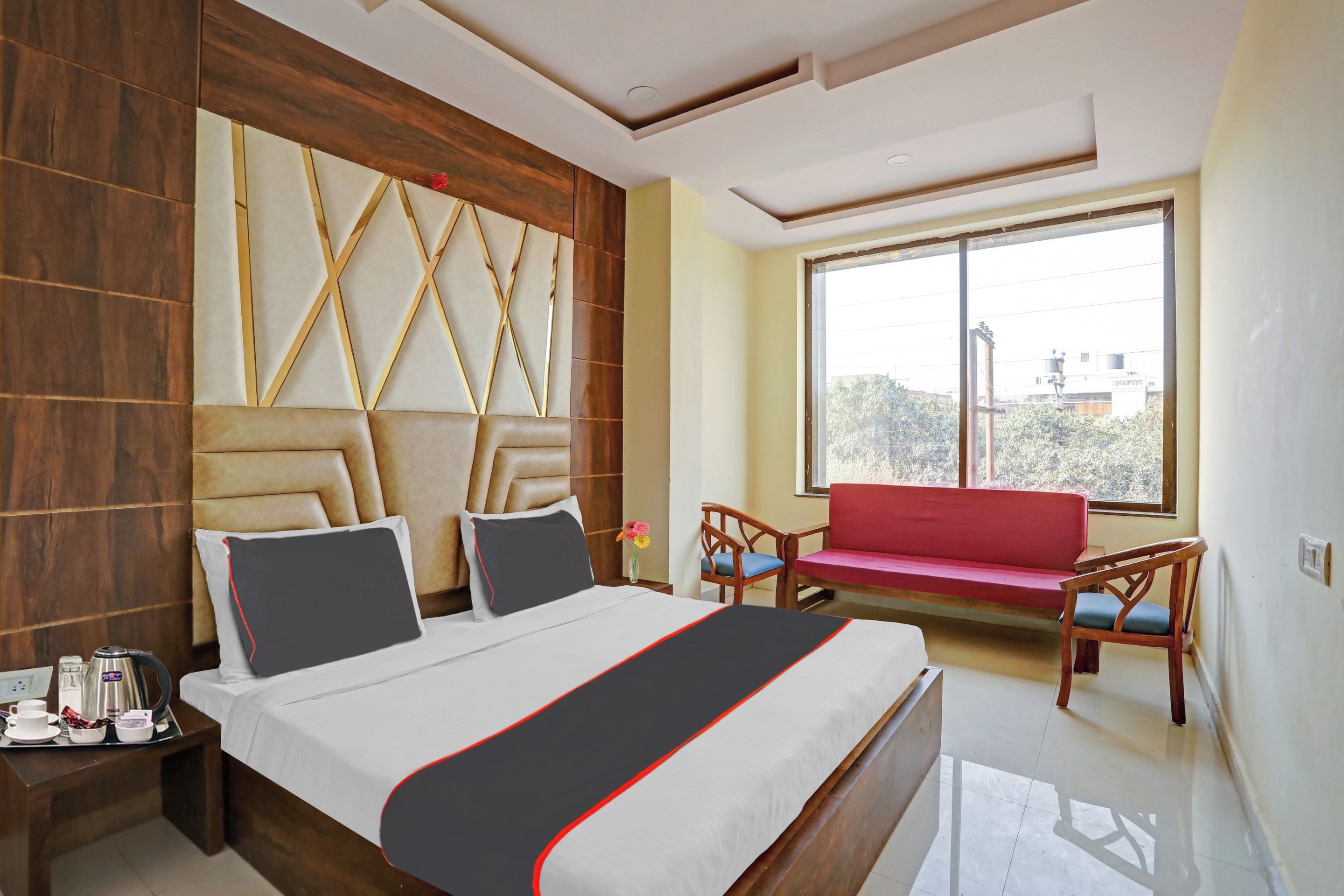 A hotel room with a bed, bedside table, a red sofa, and a window with a view.