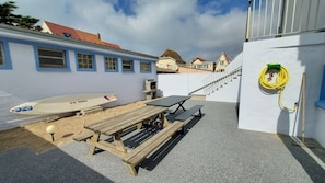 Eine Terrasse direkt am Strand mit Tisch und Bänken, einem Surfbrett und einem Gebäude mit blauen Verzierungen.