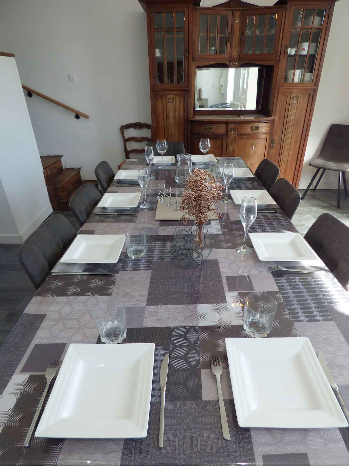 A dining table set for six with white plates, cutlery, and glasses, flanked by dark leather chairs. A wooden cabinet with glass doors is in the background.