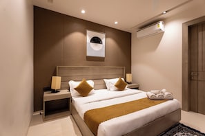 A modern hotel room with a large bed, a bedside table, a lamp, a wall-mounted air conditioner, and a door. - The Lodgers Vertex 1 BHK Apartments (Gurugram)