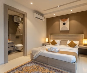 A modern hotel room with a large bed, bedside lamps, a decorative wall piece, and a bathroom with a shower and toilet. - The Lodgers Vertex 1 BHK Apartments (Gurugram)