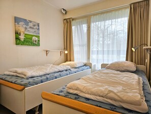 A room with two beds, a bedside lamp, a wall-mounted light, and a window with curtains. - Ok23 Oostkapelle (Oostkapelle)