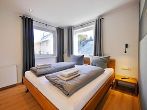 A modern bedroom with a large bed, two windows with curtains, a bedside table, and a radiator. - Mountain Magic in the Center (Willingen)