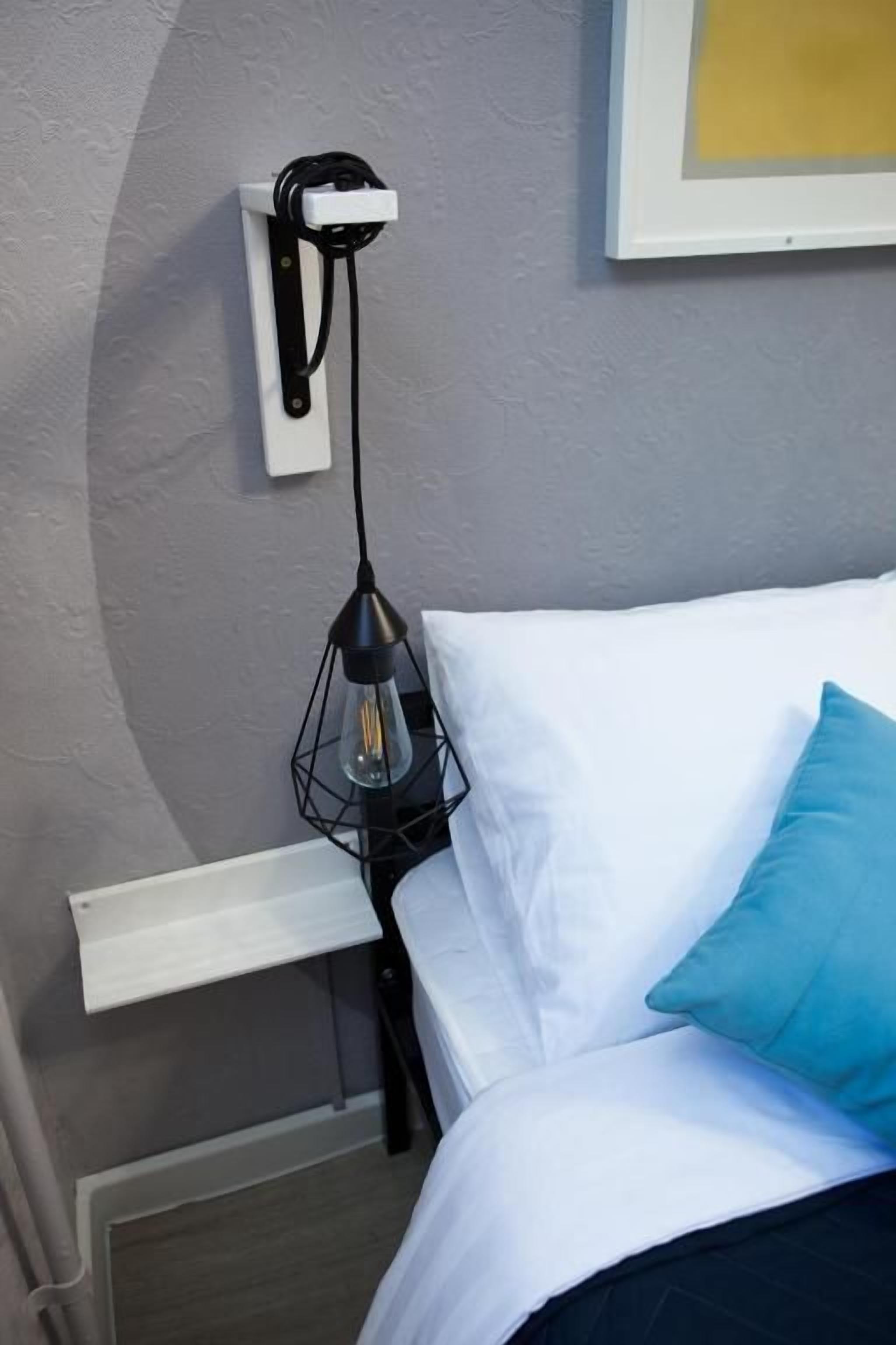 A bed with white bedding and a teal pillow, a bedside table with a lamp, and a wall-mounted power outlet.