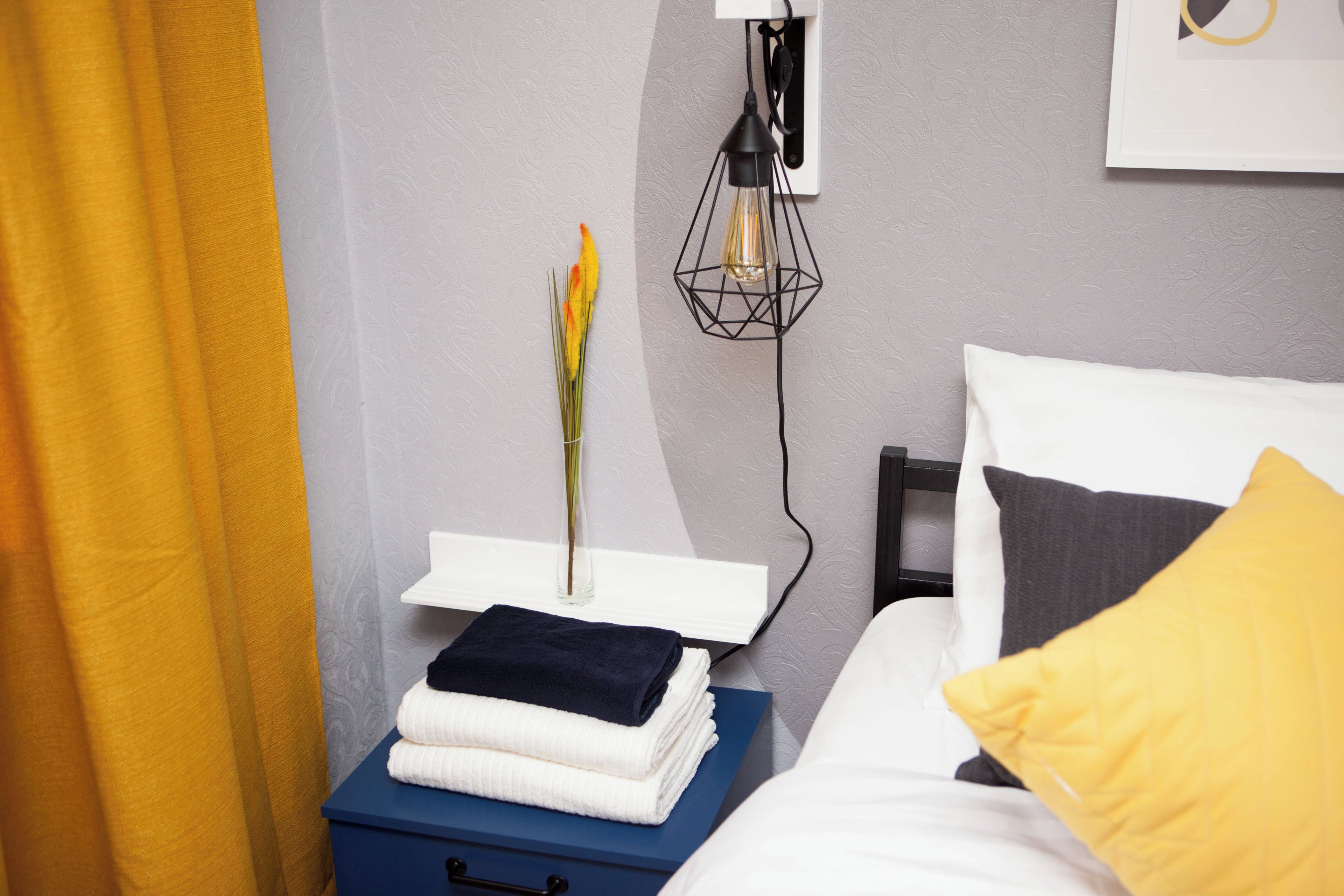 Una cama bien tendida con un almohadón blanco y uno amarillo, una mesita de noche con toallas dobladas, un florero con una flor y una lámpara de pared.