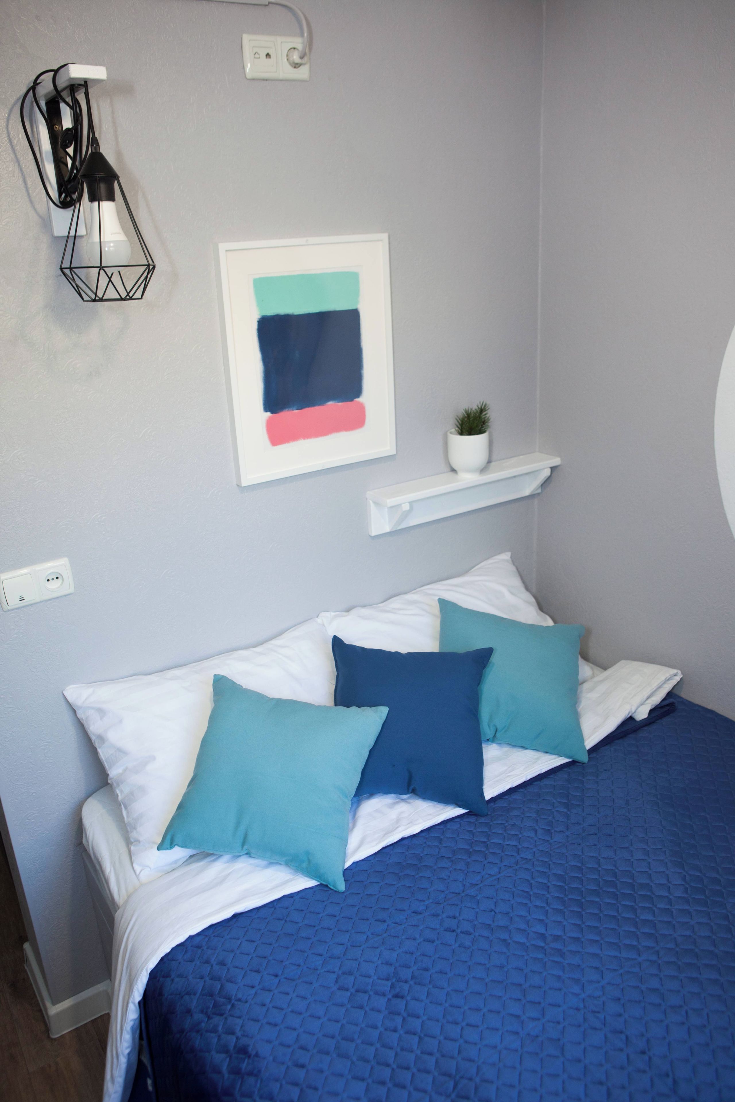 A bedroom with a bed, pillows, a wall-mounted light fixture, a framed abstract painting, and a small potted plant on a shelf.