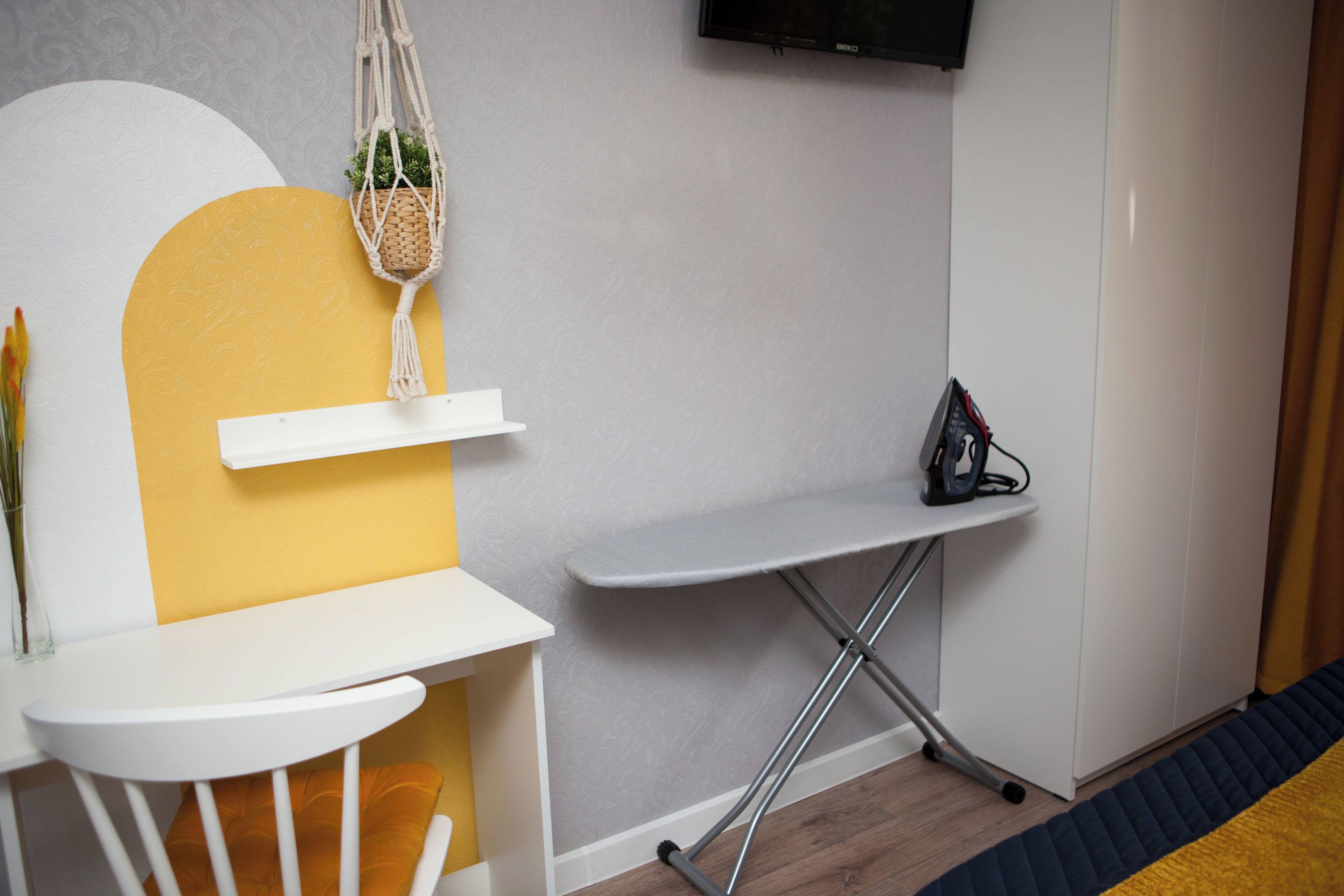 A compact room with a white and yellow wall, a small white table, a white chair, a grey foldable table, a macrame hanging plant, and a black electronic device on a grey stand.