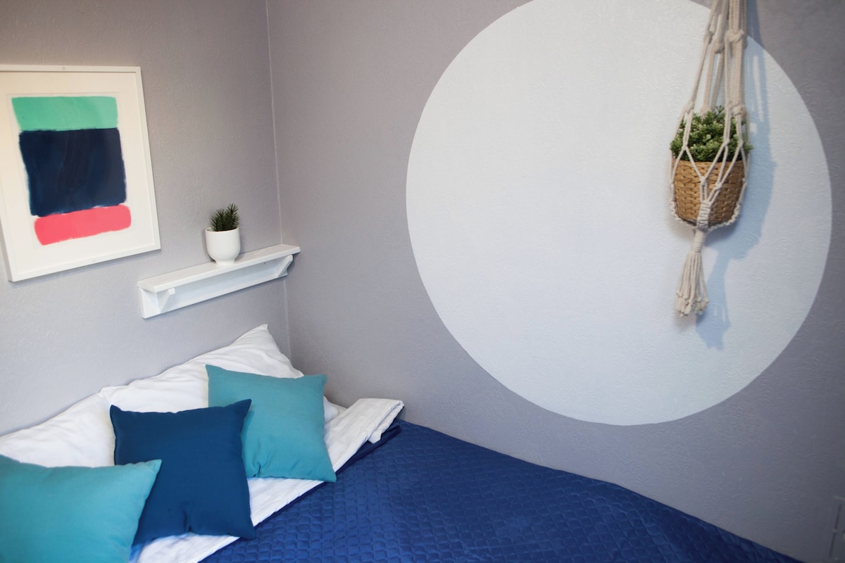A bedroom with a bed, pillows, a wall-mounted shelf with a plant, and a circular wall design.