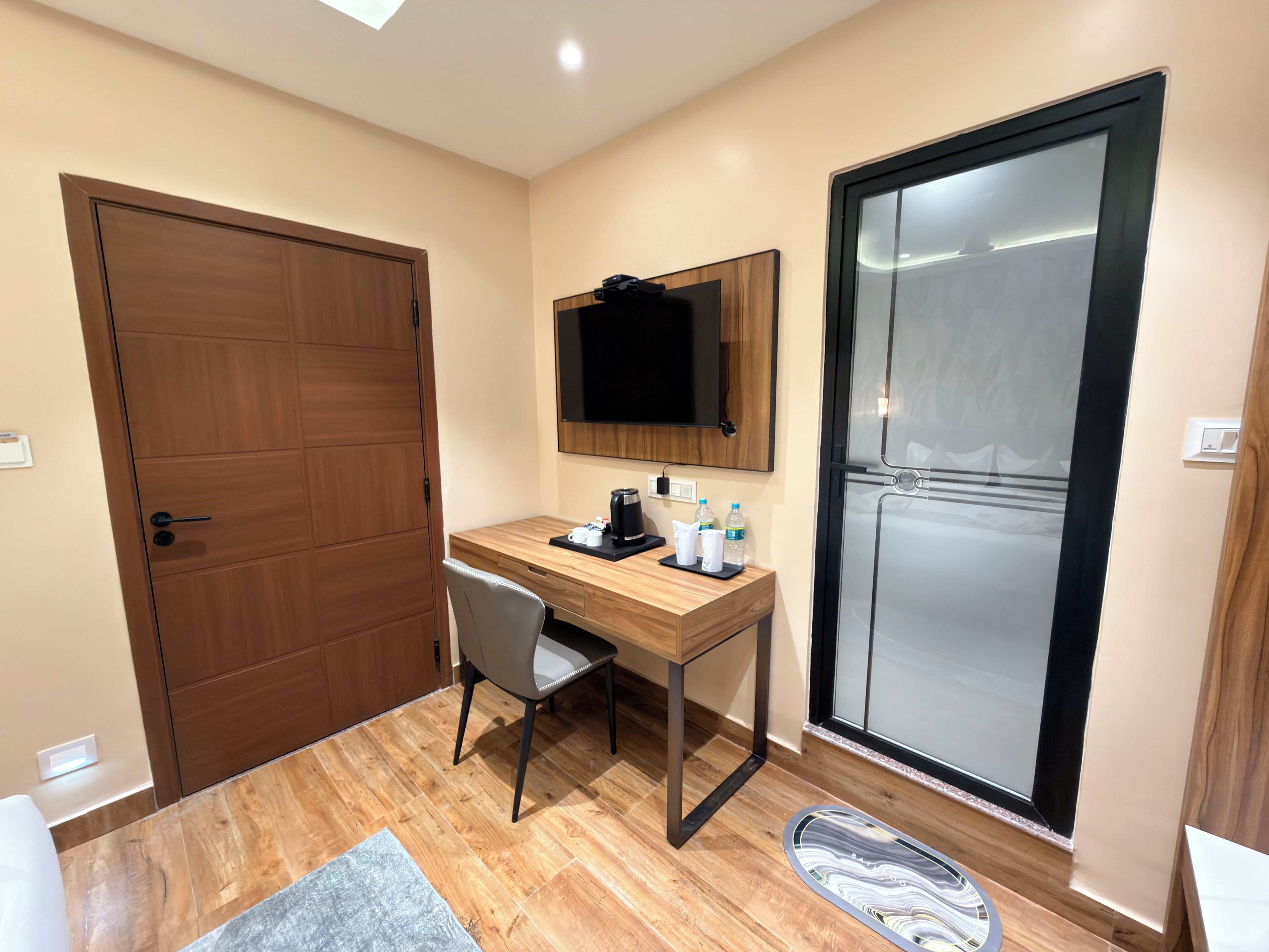 A hotel room with a wooden desk, a chair, a flat-screen TV mounted on the wall, a sliding glass door, and a small rug on the floor.