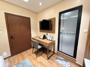 A hotel room with a wooden desk, a chair, a flat-screen TV mounted on the wall, a sliding glass door, and a small rug on the floor.