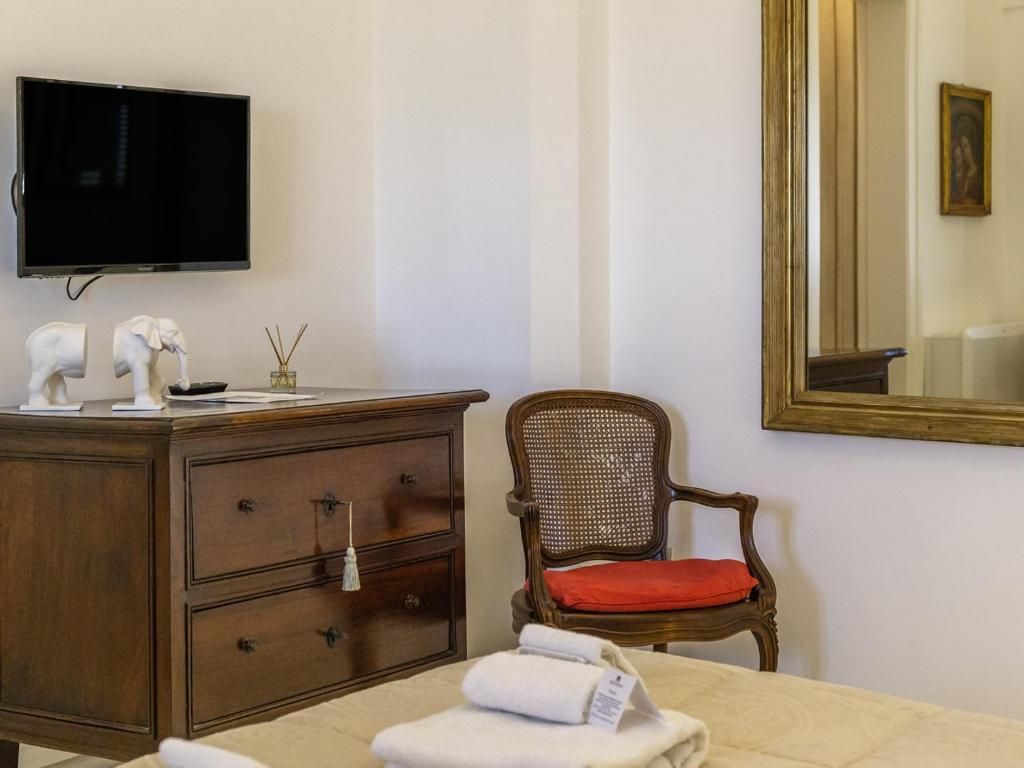 Quarto de hotel com televisão na parede, cómoda de madeira com gavetas, cadeira de vime com almofada vermelha e cama com toalhas dobradas.