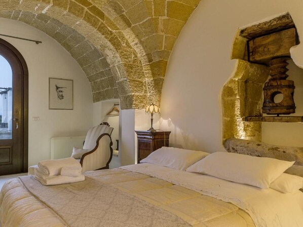 A bedroom with a stone arch ceiling, a bed with white linens, a wooden headboard, a nightstand with a lamp, and a framed picture on the wall.
