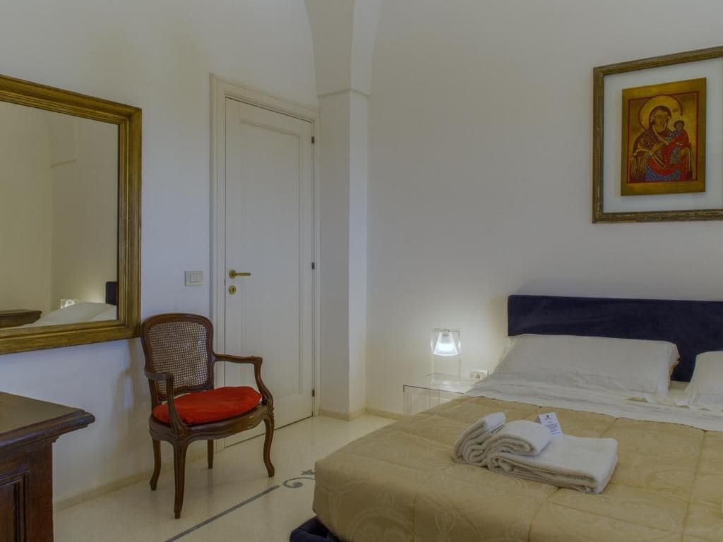 Habitación de hotel con una cama, una silla, una mesita de noche, una lámpara e ícono religioso enmarcado.