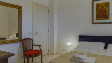 Habitación de hotel con una cama, una silla, una mesita de noche, una lámpara e ícono religioso enmarcado.