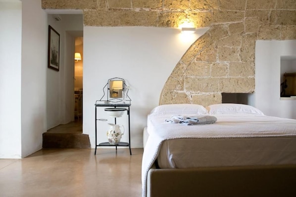 A bedroom with a stone wall, a bed with white linens, a bedside table with a lamp, and a small decorative item on the bed. - TenutaYala - Residenza Agrituristica (Parabita)