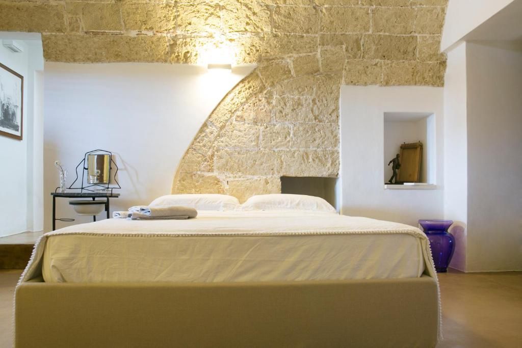 Un dormitorio con pared de piedra, una cama, una mesita de noche con lámpara y un ventanal pequeño.