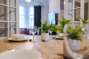 Una mesa de comedor con platos, tazones y copas de vino, con una planta en maceta y un televisor al fondo.