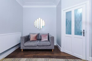Un salón moderno con un sofá gris, dos cojines, una alfombra estampada, un radiador blanco y una puerta de cristal con diseño geométrico.