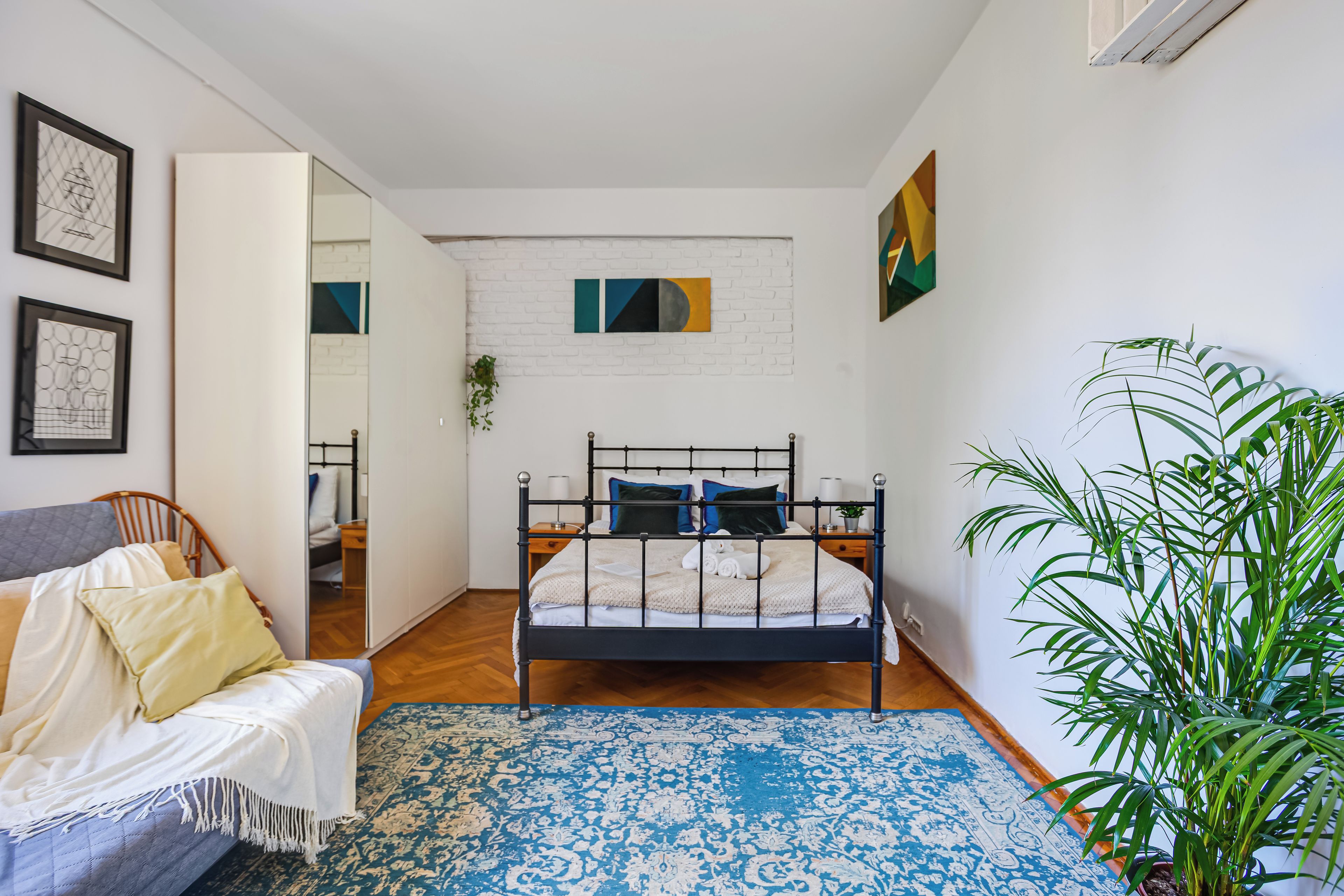 Un dormitorio con cama, sofá, alfombra estampada azul, planta en maceta y obras de arte en las paredes.