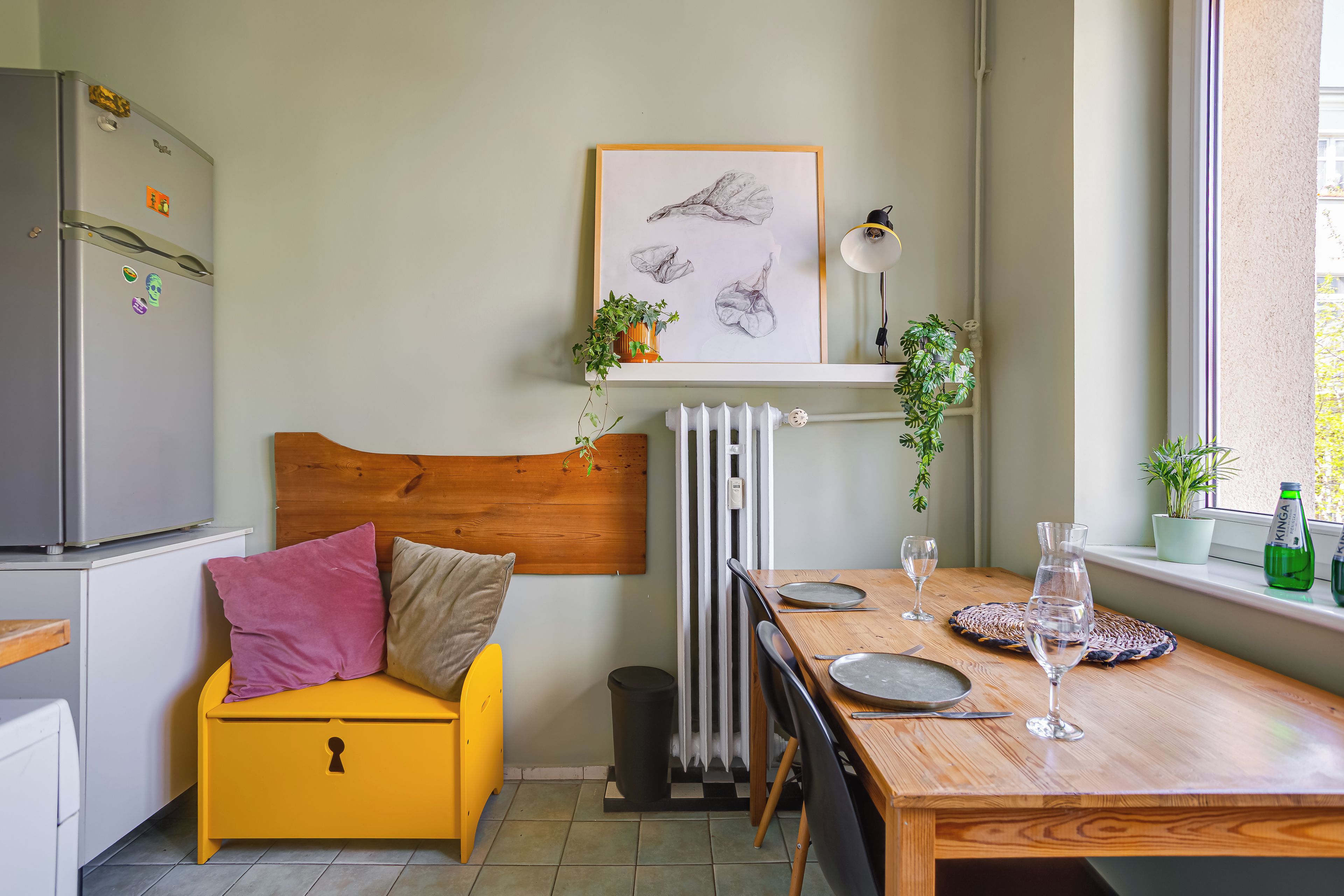 A compact living space with a bed, a dining table, a radiator, and a window with plants.