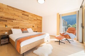 A modern bedroom with a wooden headboard, a large bed, a nightstand, a fluffy white rug, and a view of the outdoors through a glass door. - Helium—167 m² Duplex | New Construction | Terrace | View of Mont Blanc (Chamonix-Mont-Blanc)