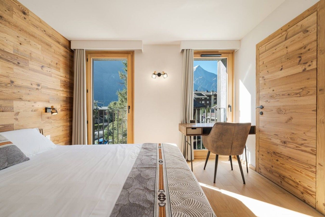 Un dormitorio con cama, escritorio y silla, paredes de madera y vistas a las montañas.