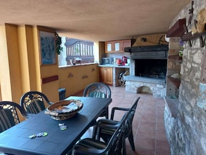 A dining area with a stone fireplace labeled 'Rustico,' a kitchen with cabinets and a microwave, and a window with a view of a fence. - Rustico La Fontana - Apt. Mimosa (Tignale)