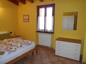 A bedroom with a bed, a nightstand, a dresser, a window with curtains, a radiator, and a framed picture on the wall. - Rustico La Fontana - Apt. Mimosa (Tignale)