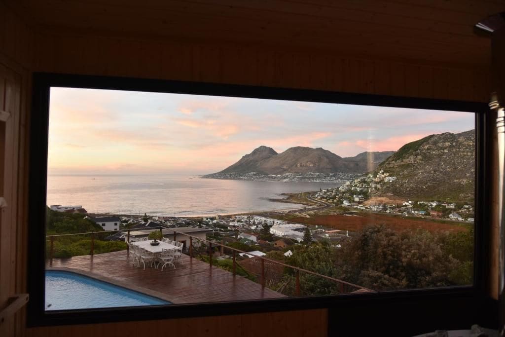 A panoramic view from a room with a large window overlooking a coastal landscape, including a body of water and distant mountains.