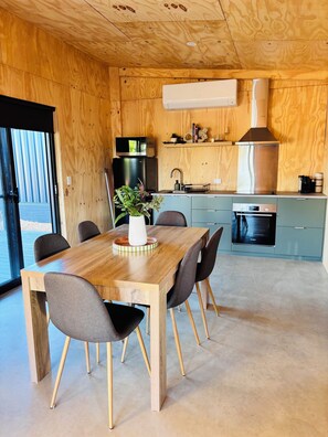 A wooden interior with a dining table, chairs, a kitchen area with appliances, and a large window. - Red Earth Retreat - raw,natural timber home (Kalbarri)