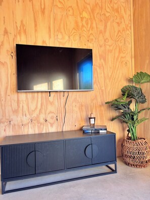 A flat-screen TV mounted on a wooden wall, a dark-colored cabinet with circular cutouts, and a potted plant with large leaves. - Red Earth Retreat - raw,natural timber home (Kalbarri)