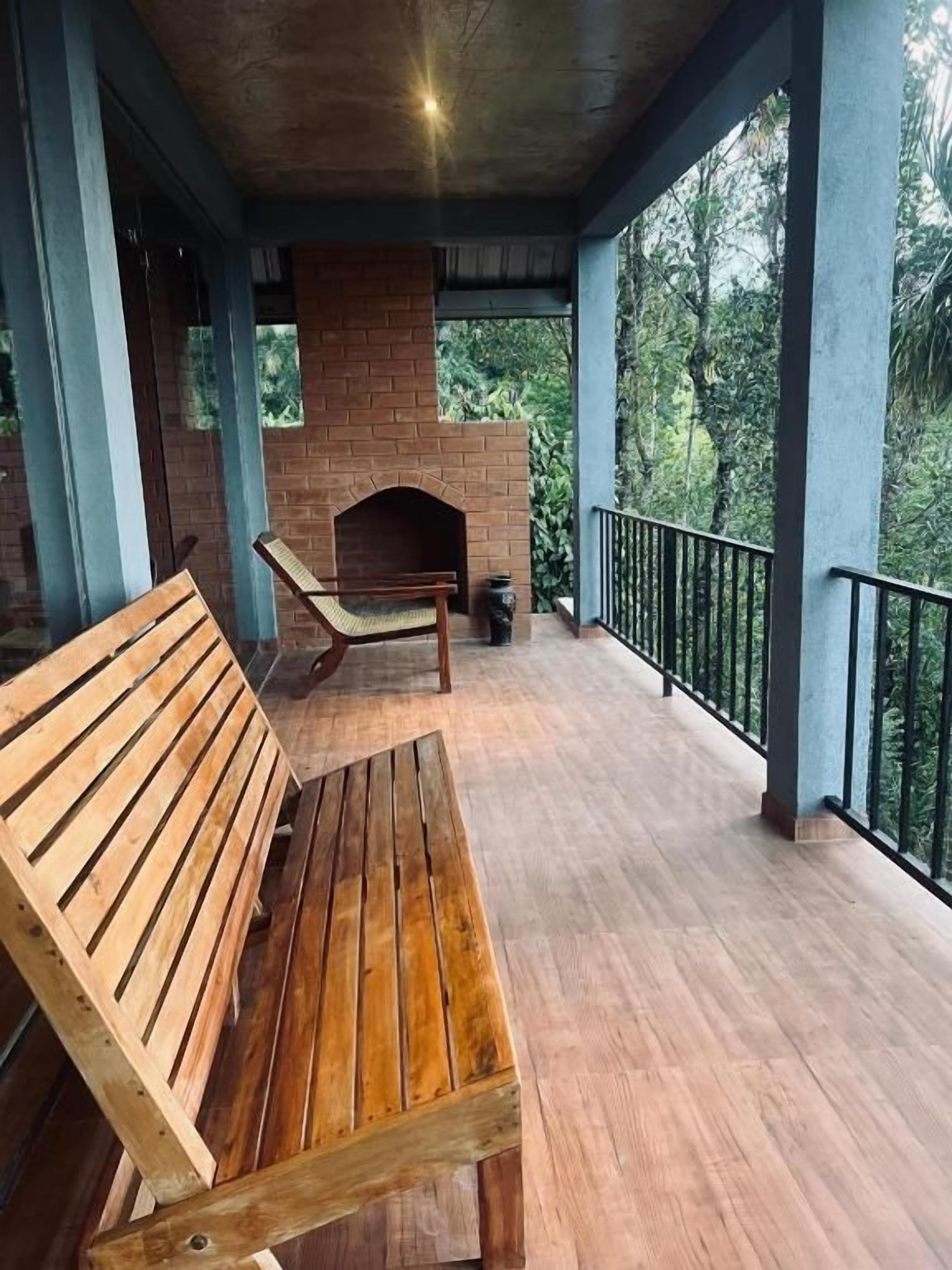 A wooden bench and a brick fireplace on a covered porch with a railing, surrounded by greenery.