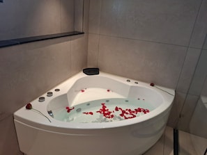 A white bathtub with rose petals in water, a black bath mat, and a rose on the edge. - SRS Elegance (Bengaluru)