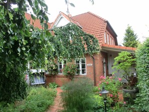 A brick house with a red-tiled roof, a garden with various plants and flowers, and a visible street sign. - Holiday Apartment With 1 Bedroom (Steinbergkirche)
