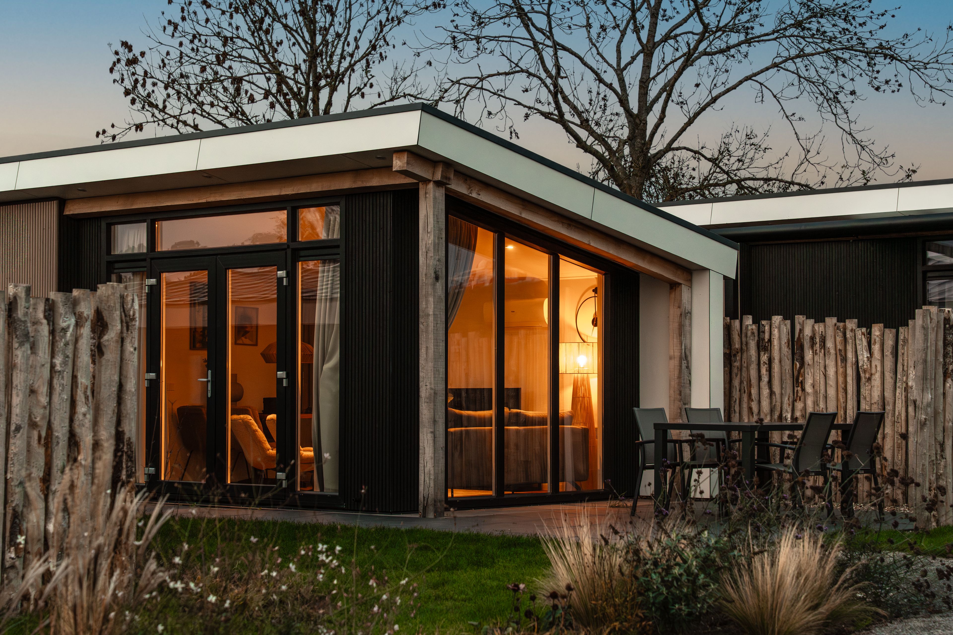 Una casa moderna con un gran ventanal que da a un espacio interior, rodeada de una valla de madera y un césped.