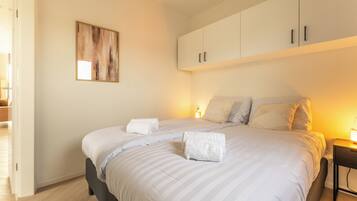A neatly made bed with white linens and two towels, a bedside table with a lamp, and a painting on the wall.