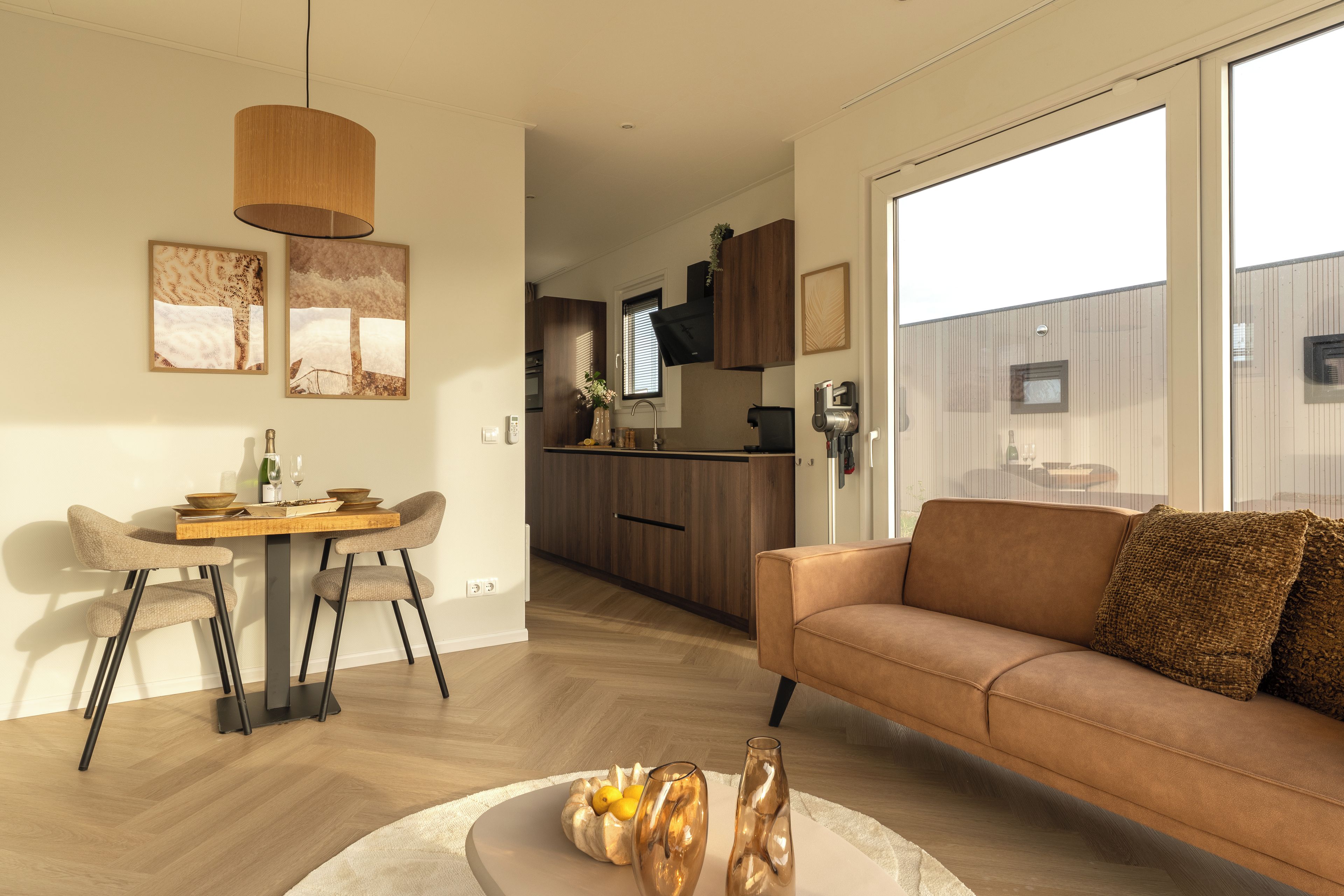 Een moderne woonkamer met een bank, een eethoek met stoelen, een keuken op de achtergrond en grote ramen.