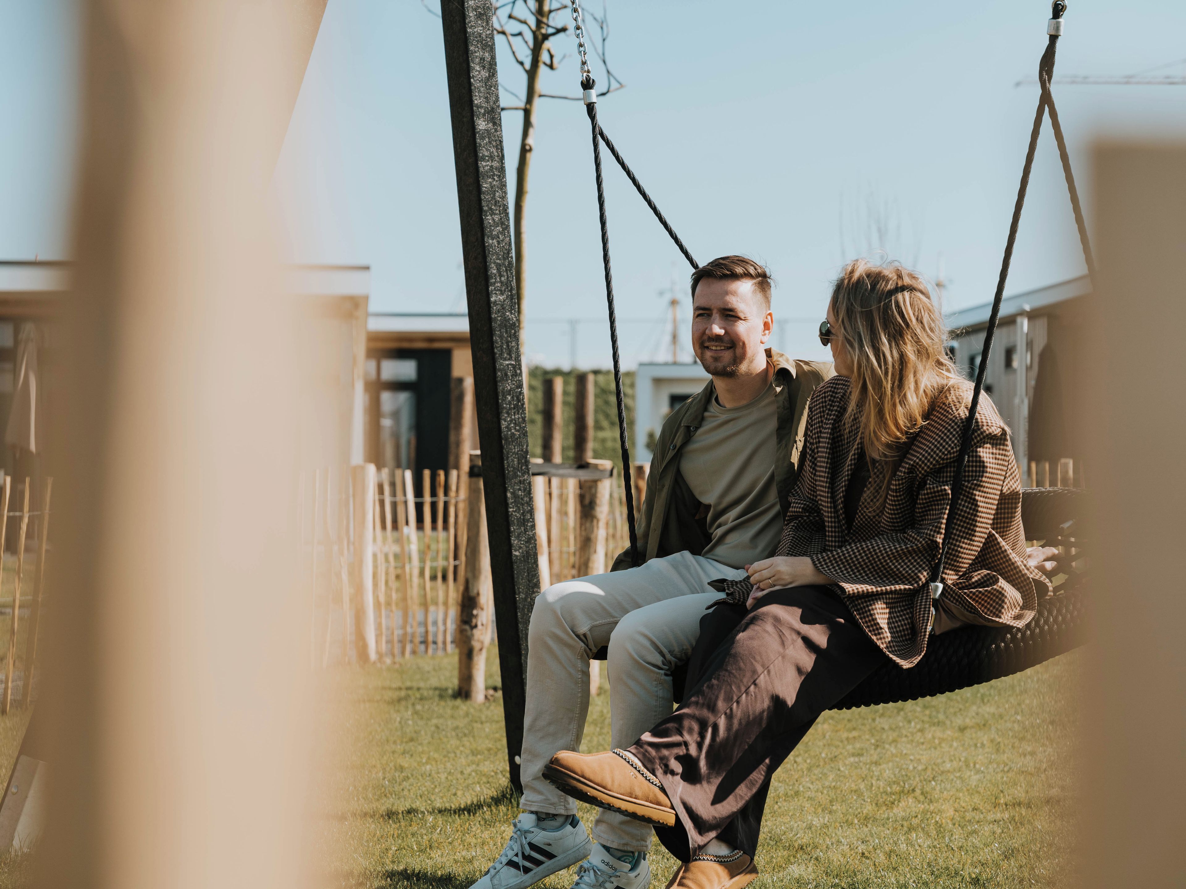 A couple sitting on a swing in a grassy outdoor area with a wooden fence and buildings in the background.