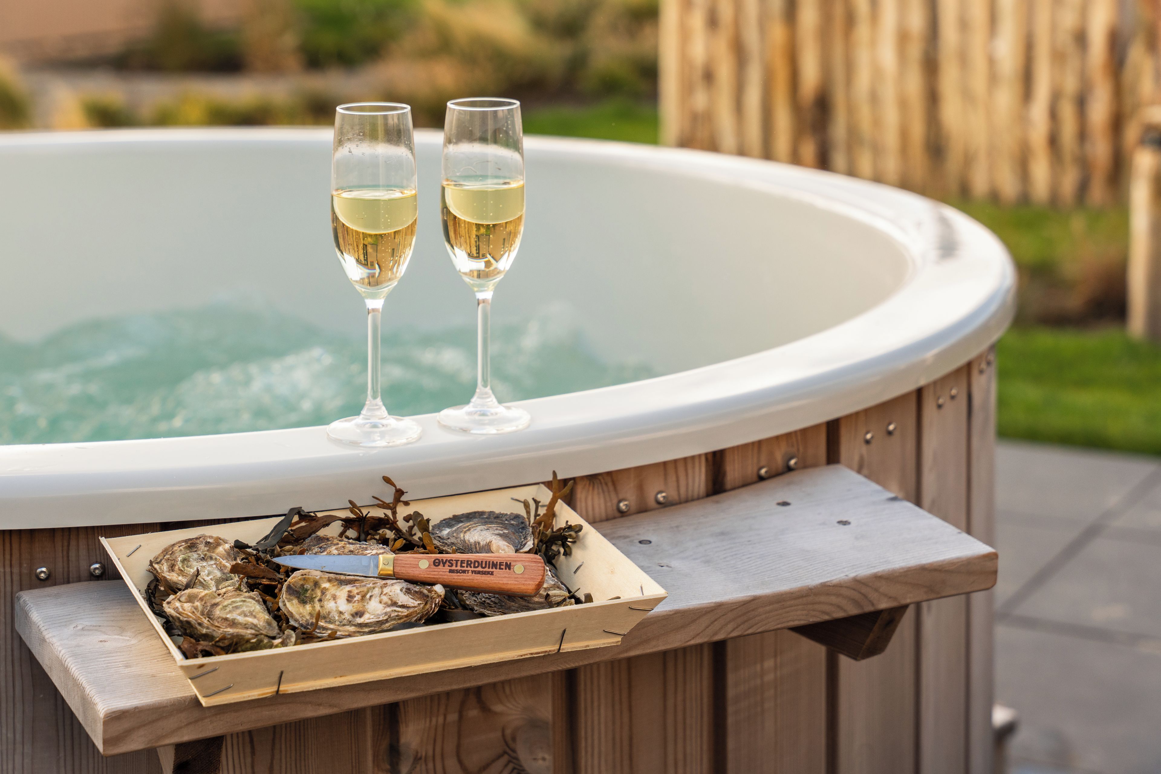A hot tub with two glasses of champagne and a wooden box containing oysters and a knife.
