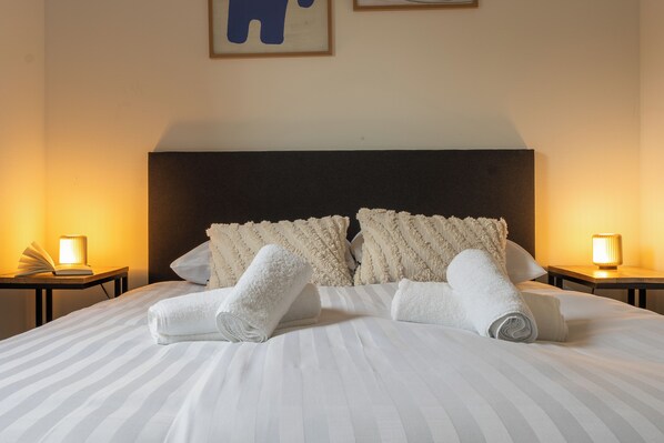 Un letto rifatto con cura, asciugamani arrotolati, una coperta a righe e due abat-jour su comodini in legno.