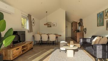 Una sala de estar moderna con televisor, muebles de madera, un comedor con sillas y una cocina al fondo.