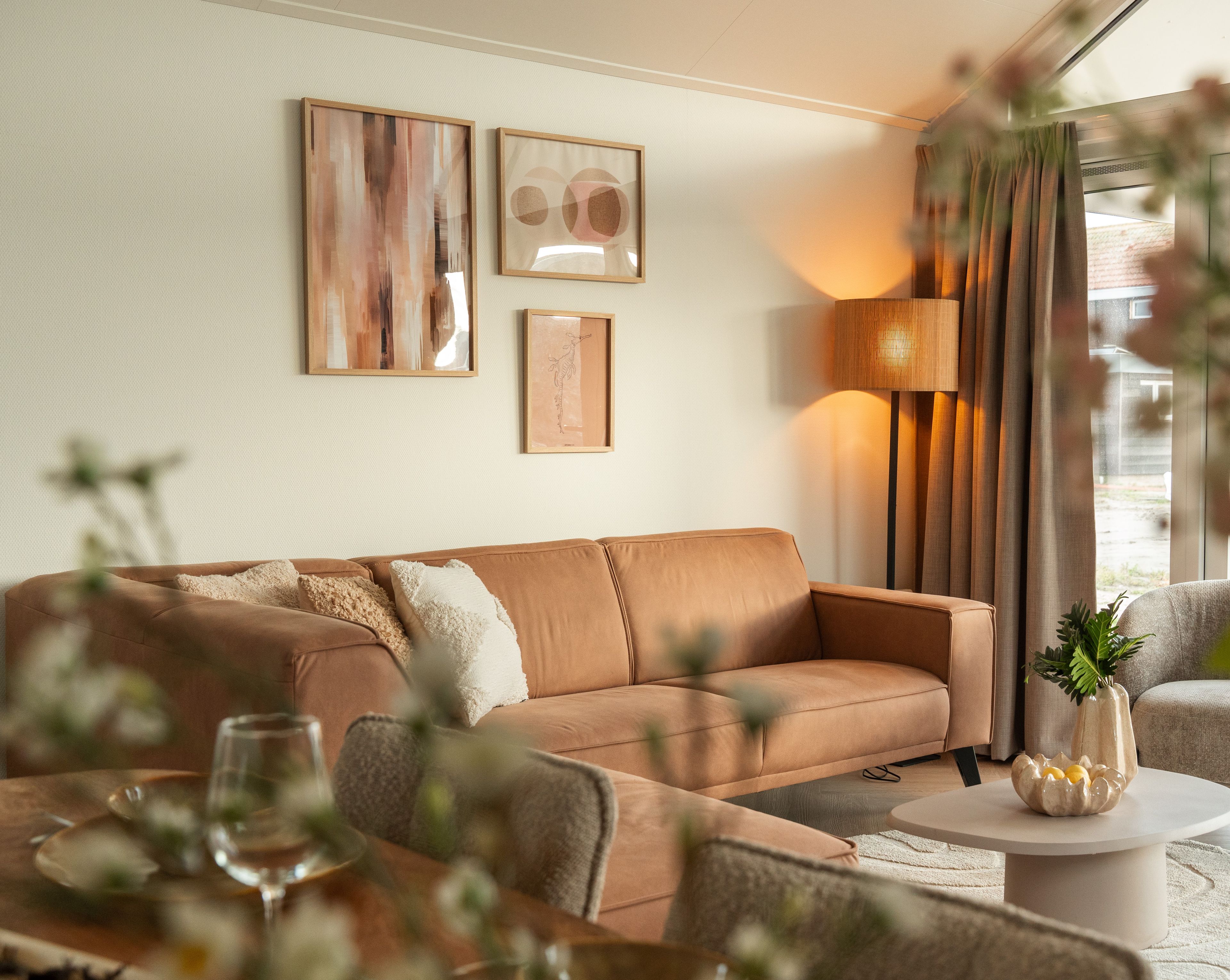 Un salon moderne avec un canapé en cuir marron, une table basse ronde et une table d’appoint sur laquelle est posé un vase. Deux œuvres d’art encadrées ornent le mur, et une lampe sur pied se trouve dans un coin de la pièce.