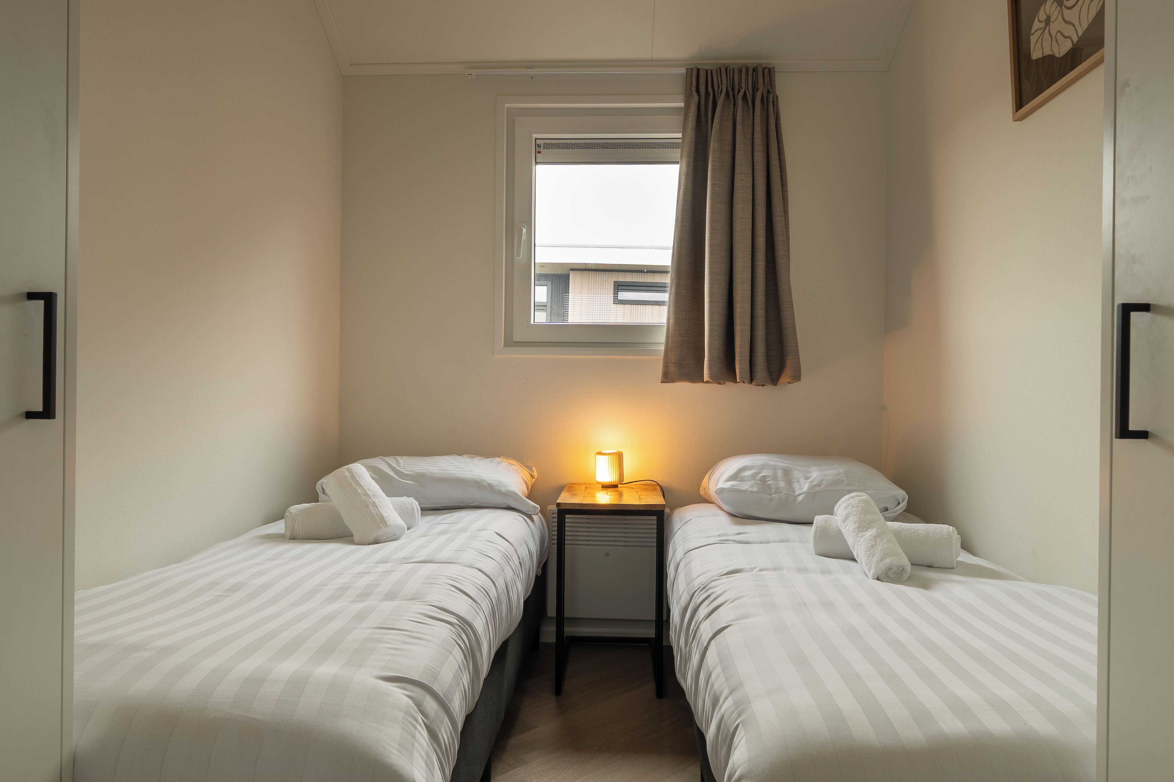 Une petite chambre bien éclairée, avec deux lits simples, une table de chevet avec une lampe et une fenêtre avec des rideaux.