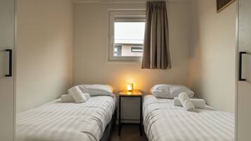 Une petite chambre bien éclairée, avec deux lits simples, une table de chevet avec une lampe et une fenêtre avec des rideaux.
