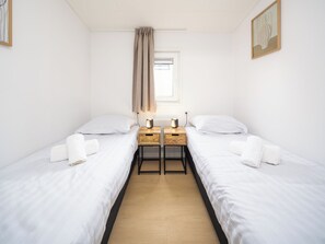 Een hotelkamer met twee eenpersoonsbedden, een tafeltje met lampen, een raam met gordijnen en een schilderij aan de muur.